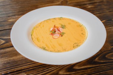 Balkabağı kremalı çorba pastırma dilimleri ve turpla süslenmiş tahta bir masada beyaz bir tabakta..