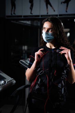 Sağlık ekibi ve sağlık maskeli bir kız spor salonunda. Coronavirus covid-19 'dan koruma. Karantina döneminde elektriksel kas uyarma giysisi sporu eğitimi