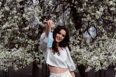 Coronavirus covid-19 periyodundan sonra tıbbi maske takan bir kız. Bahçedeki beyaz ağaçların yanında poz veren manken.