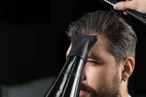 Barber dries the hair of a handsome bearded man after a fashionable haircut. The work of a hairdresser during the quarantine period coronavirus covid-19