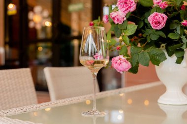 Bir bardak beyaz şarap arka plandaki güllere karşı beyaz vazoda aydınlık masada.