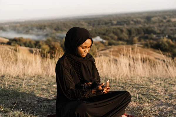 Salah. African woman in black robe sitting on the carpet and pray in god. Islamic religion.