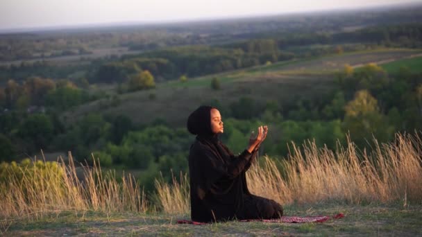 4k Salah. Afrykanka w czarnym szlafroku siedzi na dywanie i modli się w Bogu. Religia islamska — Wideo stockowe