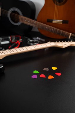 Arka planda gitar ve aletleri olan gitar seçimleri. Gitar ve tamir aletleriyle siyah bir yüzey üzerinde renkli gitar penaları bulunuyor. Masanın üzerinde parlak renkte çoklu gitar penaları