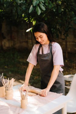 Çamur, çömlek ve seramik sanat için iğne şekilli malzemeyle yuvarlanıyor. Masanın üzerinde yuvarlanan bir çivi düzleştirici kil var. Çamur, seramik sanat için zemin hazırlıyor.