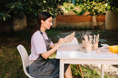 Genç bir kadın çömlekçilik yeteneklerini sergilemek için kilin içine ince yaprak desenleri oyuyor. Esnaf, üzerinde ayrıntılı çizgiler olan bir kil yaprak oyar. Çamur Yaprağı Oyan Kadın