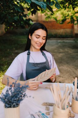 Genç bir kadın çömlekçilik yeteneklerini sergilemek için kilin içine ince yaprak desenleri oyuyor. Esnaf, üzerinde ayrıntılı çizgiler olan bir kil yaprak oyar. Çamur Yaprağı Oyan Kadın