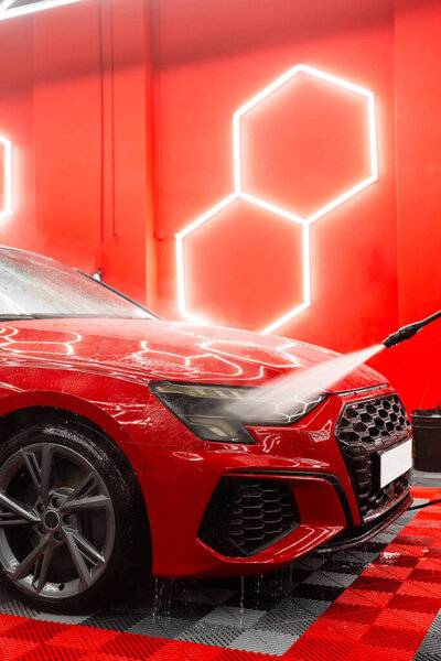 High pressure car rinse removes dirt, foam, and road grime from body panels and headlights of red sports car. Paint safe car rinse with high pressure water removes debris, soap residue, and dirt