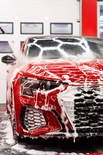 Red sports car detailing rinses thick snow foam with high-pressure water, removing dirt and leaving a streak-free, glossy finish. High-pressure water to flush foam. Paint-safe foam rinse removes foam