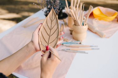 Genç bir kadın çömlekçilik yeteneklerini sergilemek için kilin içine ince yaprak desenleri oyuyor. Esnaf, üzerinde ayrıntılı çizgiler olan bir kil yaprak oyar. Çamur Yaprağı Oyan Kadın