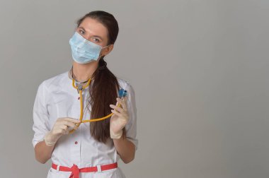 Genç bir doktor, beyaz önlüklü, elinde steteskop, gri arka planda tıbbi maskeli bir kadın.