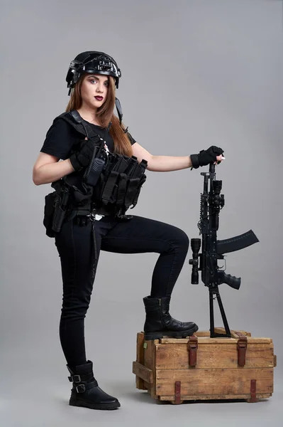 Une jeune femme avec une mitrailleuse appuyée sur une boîte de munitions sur un fond gris — Photo