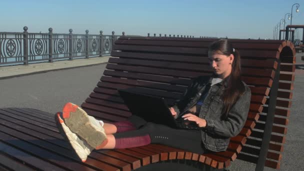 Mujer joven sentada en un banco con un portátil y trabajando en un día soleado de verano — Vídeos de Stock