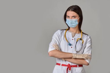 Beyaz önlüklü, steteskoplu ve gri izole arka planda tıbbi maskesi olan genç bir kadın doktor. Boşluğu kopyala