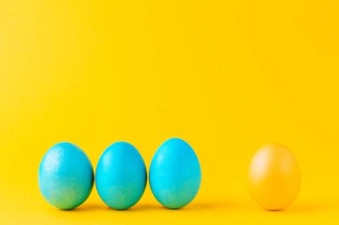Tek sarı yumurtaya karşı üç mavi yumurta. Sarı arka planda renkli Paskalya yumurtaları. Farklılık kavramı.