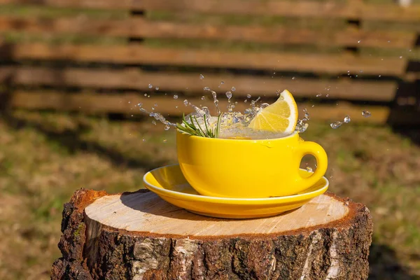 Splash of tea in yellow cup with lemon on wooden log in the yard. Fresh summer drink with flying drops in bright sunlight outdoors.