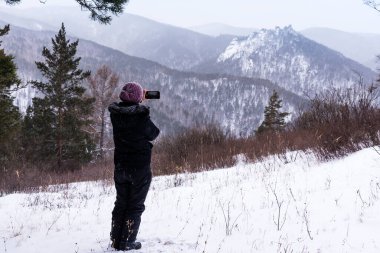 Bir kadın, kışın kayalık bir tepenin cep telefonunda fotoğraf çeker. Güzel manzara, yürüyüş, açık hava aktiviteleri, turizm.