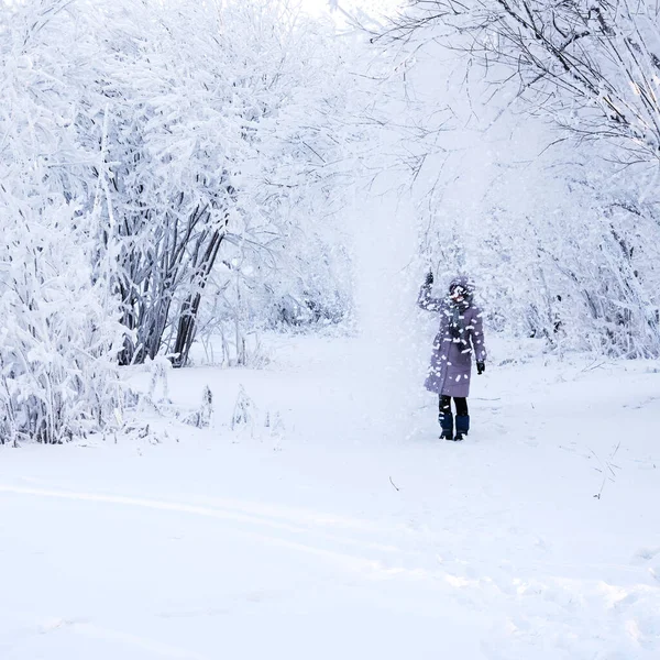 Woman standing under falling snow in winter forest. Carefree woman enjoying snow and winter time,  playing with snow. Winter leisure concept.
