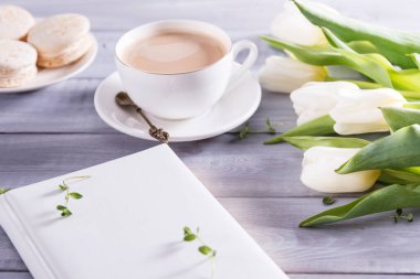 Bir fincan kahve, badem kurabiyesi ve beyaz ahşap arka planda bir buket beyaz lale. Düz uzanış, üst görünüm kadın masası, çalışma odası ve defter. Güzellik günlüğü konsepti