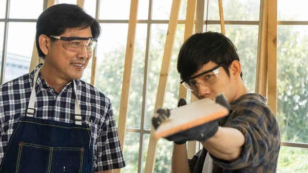 Two Asian carpenters working together in workspace. mature adult father and growing son preparing wood and tools to repair house. young man learning carpentry and handicrafts with senior master.