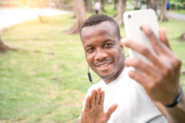 Genç bir Afrikalı Amerikalı parkta akıllı telefonuyla konuşuyor. Genç siyah erkek sporcu yeşil ormanda antrenman yaptıktan sonra rahatlıyor. yakışıklı sporcu selfie ve vlog egzersiz yaparken yayınlanıyor.