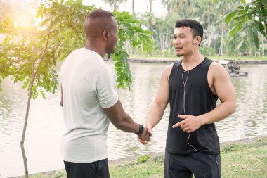 Asyalı ve Afrikalı kaslı genç erkek arkadaşlar el sıkışır, selamlaşır, parkta gülümser. Erkek sporcu ve antrenör dışarıda antrenman yaptıktan sonra rahatlar. İki sporcu göl kenarında müzik ve egzersiz dinlerler..