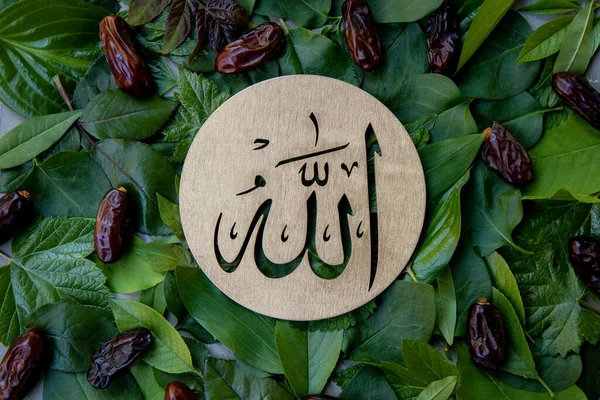 Name of Allah in arabic on gold wood, green leaves and dates fruit background. Calligraphy means the God Al Mighty of Islam Religion.