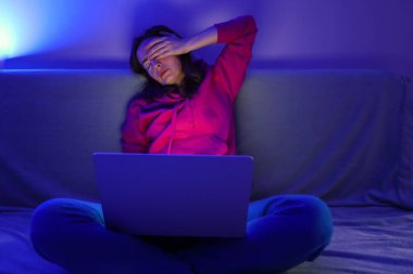 Yorgun kadın gece mavi ışıkla dizüstü bilgisayarda çalışırken baş ağrısı ve yanma çekiyor..