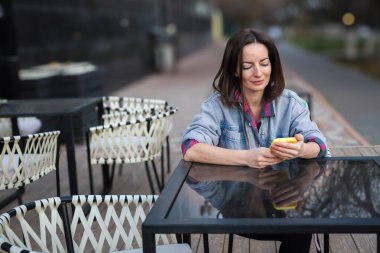 40 'lı yaşlarda güzel bir kadının portresi. Elinde sarı çantalı bir cep telefonu var. Şehirdeki bir kafedeki masada oturan, mesaj yazan, internetten sohbet eden ya da mobil uygulama kullanan bir kadın. Kentsel yaşam tarzı ve teknoloji kavramı.