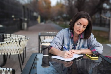 Orta yaşlı bir kadın not defterine not yazıyor ya da yapılacaklar listesi hazırlıyor. Açık hava bir şehir kafesinde bir masada otururken çalışan, ders çalışan ya da günlük tutan meşgul bir kadın. Zaman yönetimi, planlama ve organizasyon kavramı.