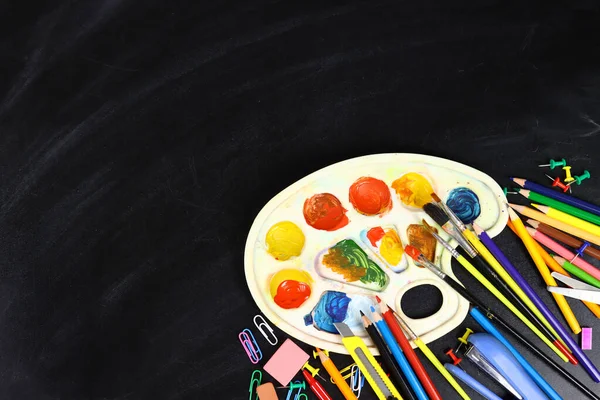 colorful pencil, paints, brushes and stationery flat lay on the blackboard, back to school concept