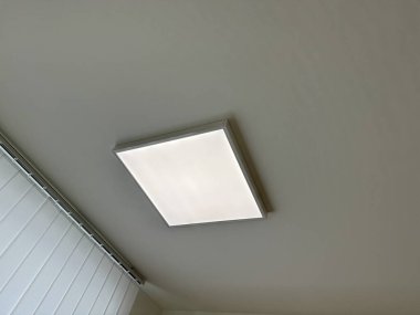 Pencere perdelerinin yanındaki beyaz tavana monte edilmiş panel aydınlatma armatürü