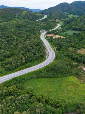 Aerial drone view of Highway 101 winding through lush green mountains between Phrae and Nan, Thailand. A scenic landscape of the road surrounded by tropical forest and a peaceful countryside 