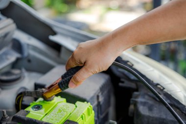 Araba aküsüne yakın bir jumper kablo kıskacını tutan bir el araç bakımını, acil durum başlatmasını ve otomotiv tamiri hazırlıklarını gösteren bir el.
