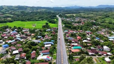 High-angle drone footage showing a long highway passing through a rural village surrounded by green fields and forested hills, capturing countryside lifestyle, transportation routes