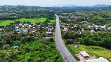 Yüksek açılı insansız hava aracı görüntüleri yeşil tarlalar ve orman tepeleriyle çevrili kırsal bir köyden geçen uzun bir otoban gösteriyor. Kırsal yaşam tarzını, ulaşım yollarını yakalıyor.