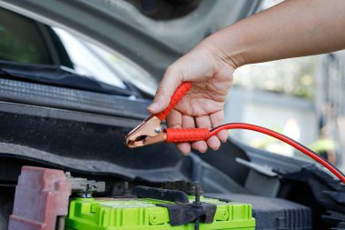 Araba aküsüne yakın bir jumper kablo kıskacını tutan bir el araç bakımını, acil durum başlatmasını ve otomotiv tamiri hazırlıklarını gösteren bir el.