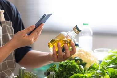 Orta yaşlı Asyalılar evde sağlıklı yiyecekler hazırlarken zeytinyağını akıllı telefonla kontrol ediyorlar. Sağlıklı yaşlanma, iyi yağ seçimi, beslenme farkındalığı ve koruyucu yaşam tarzı kavramı