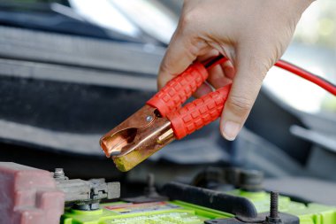 Araba aküsüne yakın bir jumper kablo kıskacını tutan bir el araç bakımını, acil durum başlatmasını ve otomotiv tamiri hazırlıklarını gösteren bir el.