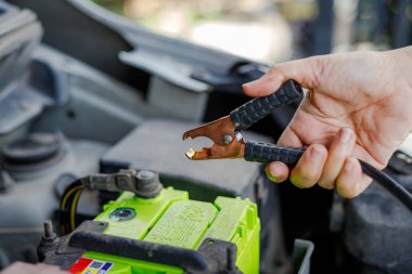 Araba aküsüne yakın bir jumper kablo kıskacını tutan bir el araç bakımını, acil durum başlatmasını ve otomotiv tamiri hazırlıklarını gösteren bir el.