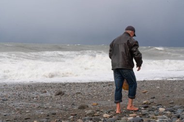 Çakıl taşlı sahilde yalın ayak yürüyen bir adam.