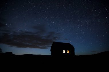 Yıldızlı gökyüzünün arka planında güzel bir köy evinin silueti. Karanlık gece. Işık ve gölge oyunu. Güzel yıldızlı gökyüzü. Evin pencerelerinde ışık var. Kırsal alan.