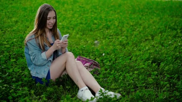 Junges schönes Mädchen sitzt auf dem Gras und zeigt in das Telefon — Stockvideo