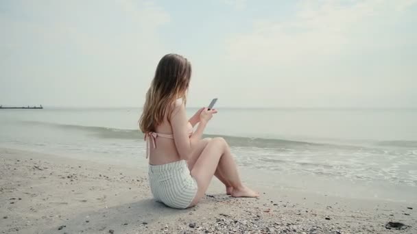 Ung kvinna med hjälp av mobiltelefon på stranden, dolly skott — Stockvideo