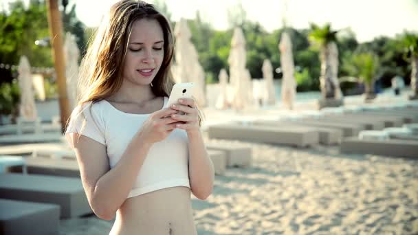 Mulher usando smartphone com fones de ouvido rindo na praia ouvindo música. Menina de biquíni usando — Vídeo de Stock