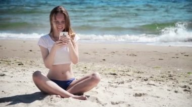 Hareket eden telefon yanında deniz ve mavi gökyüzü kum üzerinde oturan kız. İlk aşk.