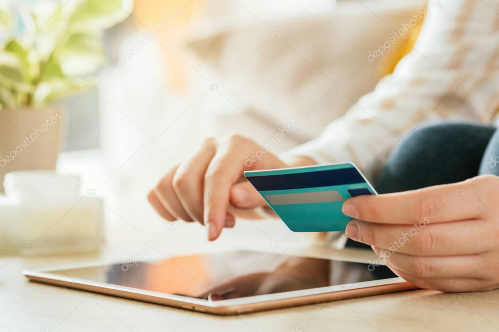 Stock photo of an unrecognizable caucasian woman shopping online with the tablet and a credit card from the couch at living room. E-commerce and buying from sofa concept