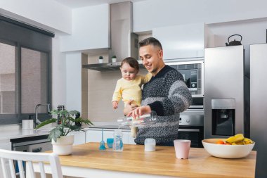 Genç bekar babanın, bebeğini mutfakta kucağında tutarken bir beslenme şişesi hazırlarken çekilmiş resmi. Erkek ve çocuk, gerçek bir baba olma anı. Bekar baba aile hayatı konsepti.