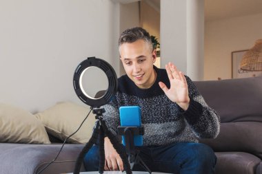 Genç adam milenyum etkileyicisinin resmi kanepeye oturmuş akıllı telefon ve ışıklarla çevrimiçi içerik kaydediyor, kaydediyor ve yaratıyor. Sosyal medya video kanalı kayıt kavramı