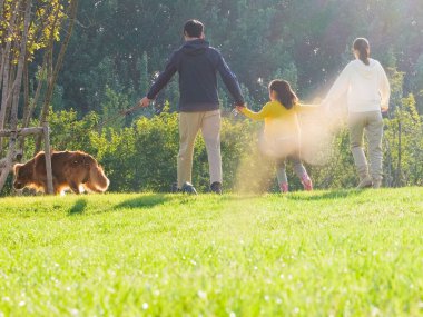 Üç kişilik mutlu bir aile ve parkta yürüyen evcil bir köpek.